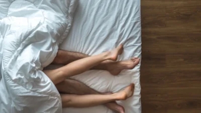 A couple is lying on a bed with white sheets, exploring questions about sex they recently googled.