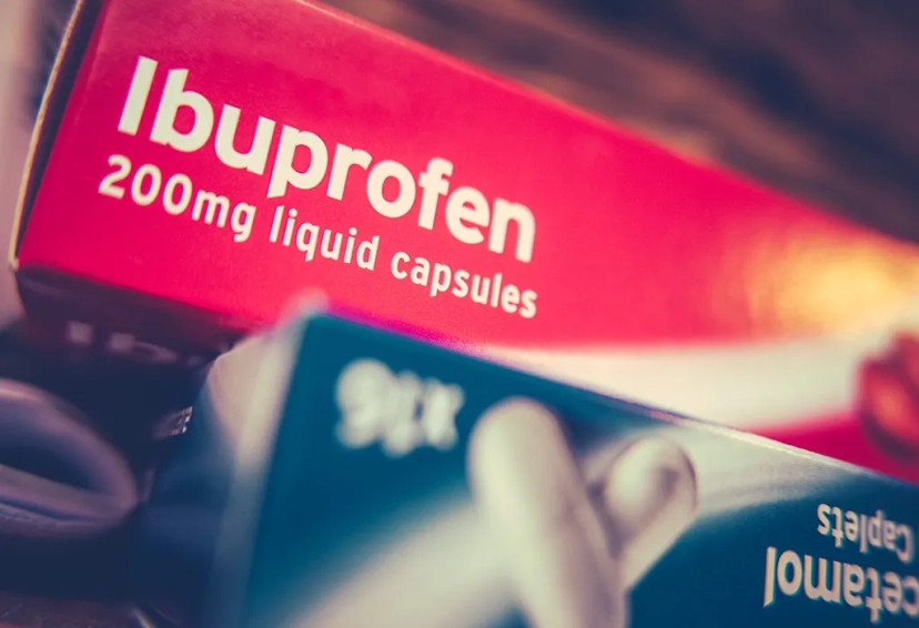 Close-up of a red box labeled "Ibuprofen 200mg liquid capsules," designed to help with stomach pain, partially overlapping a visible portion of another box of tablets in the foreground.