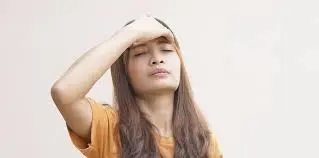 A person with long hair and wearing an orange shirt holds their forehead with eyes closed, appearing to be in discomfort, likely experiencing symptoms of hay fever.