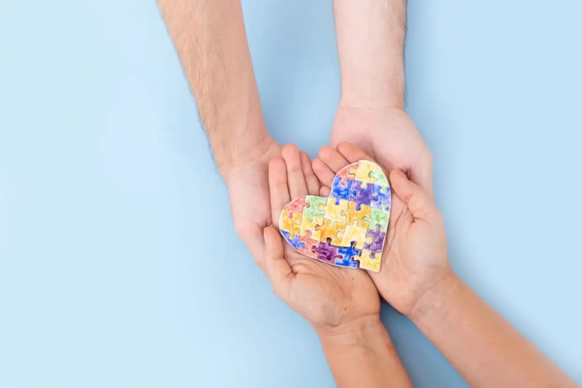 Two pairs of hands holding a heart-shaped puzzle piece with colorful patterns against a light blue background, symbolizing understanding and support for those on the autism spectrum.