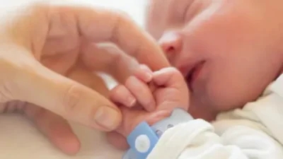 Close-up of a newborn baby holding an adult's finger. The baby, wearing a hospital identification band on its wrist, is blissfully unaware of the potential transmission risks such as group B strep infection.