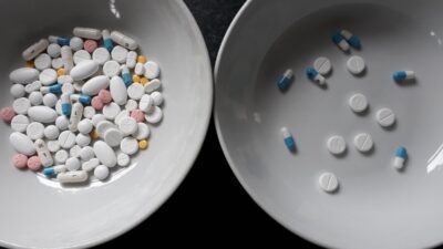 Two white bowls are displayed; the left bowl is filled with a variety of pills and tablets in different colors and shapes, while the right bowl, indicative of deprescribing efforts, contains a smaller assortment of white pills and blue-white capsules.
