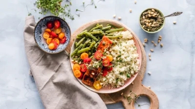 A wooden board displays a plate of quinoa, roasted vegetables, and green beans. Beside it, a bowl of cherry tomatoes and a bowl of seeds with a spoon rest on the light blue surface with a gray napkin. These foods are delicious yet considerate for those mindful of acid reflux triggers.