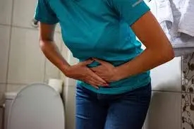 Person in a blue shirt and jeans holding their stomach, appearing to be in pain, standing near a toilet in a bathroom, possibly concerned about a UTI.