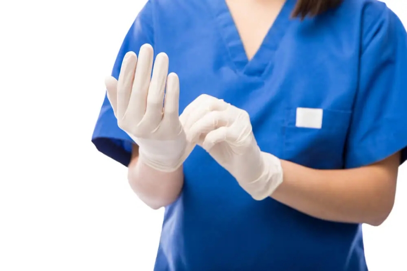 A person in blue scrubs is putting on white medical gloves, taking careful measures for infection control. Their face is not visible in the image.