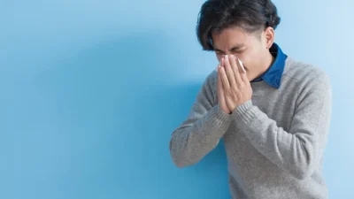 A person standing against a blue background, sneezing into a tissue while wearing a gray sweater, indicating signs of an upper respiratory tract infection.