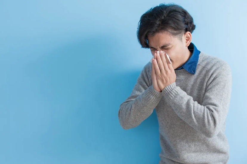 A person standing against a blue background, sneezing into a tissue while wearing a gray sweater, indicating signs of an upper respiratory tract infection.