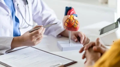 A doctor holds a pen and points at a heart model on a desk, explaining the difference between stable angina and unstable angina, with a clipboard and a patient's hands in the foreground.