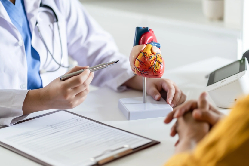 A doctor holds a pen and points at a heart model on a desk, explaining the difference between stable angina and unstable angina, with a clipboard and a patient's hands in the foreground.