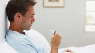 A man sits up in bed, looking at a thermometer in his hand, contemplating how to reduce his fever.