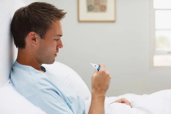 A man sits up in bed, looking at a thermometer in his hand, contemplating how to reduce his fever.