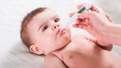 A baby lies on a white surface while an adult hand places a digital thermometer in the child's mouth to check if their temperature is too high.