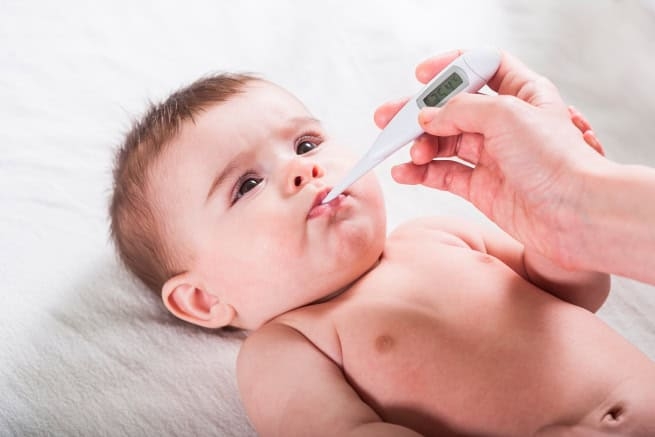 A baby lies on a white surface while an adult hand places a digital thermometer in the child's mouth to check if their temperature is too high.