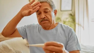 Elderly man in a light blue shirt holding a thermometer, with a concerned expression and his other hand on his forehead, contemplating the fever duration, indoors.