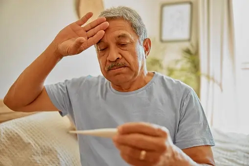 Elderly man in a light blue shirt holding a thermometer, with a concerned expression and his other hand on his forehead, contemplating the fever duration, indoors.