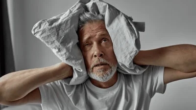 Elderly man with a distressed expression, covering his ears with a pillow, showing signs of schizophrenia