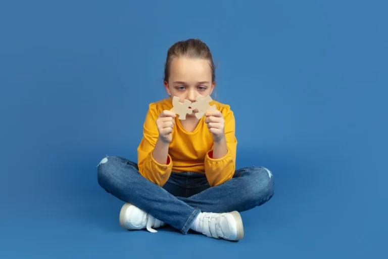 A child in a yellow long-sleeve shirt and jeans sits cross-legged against a blue background, holding two large beige puzzle pieces, symbolizing the connection between autism and genetic factors.