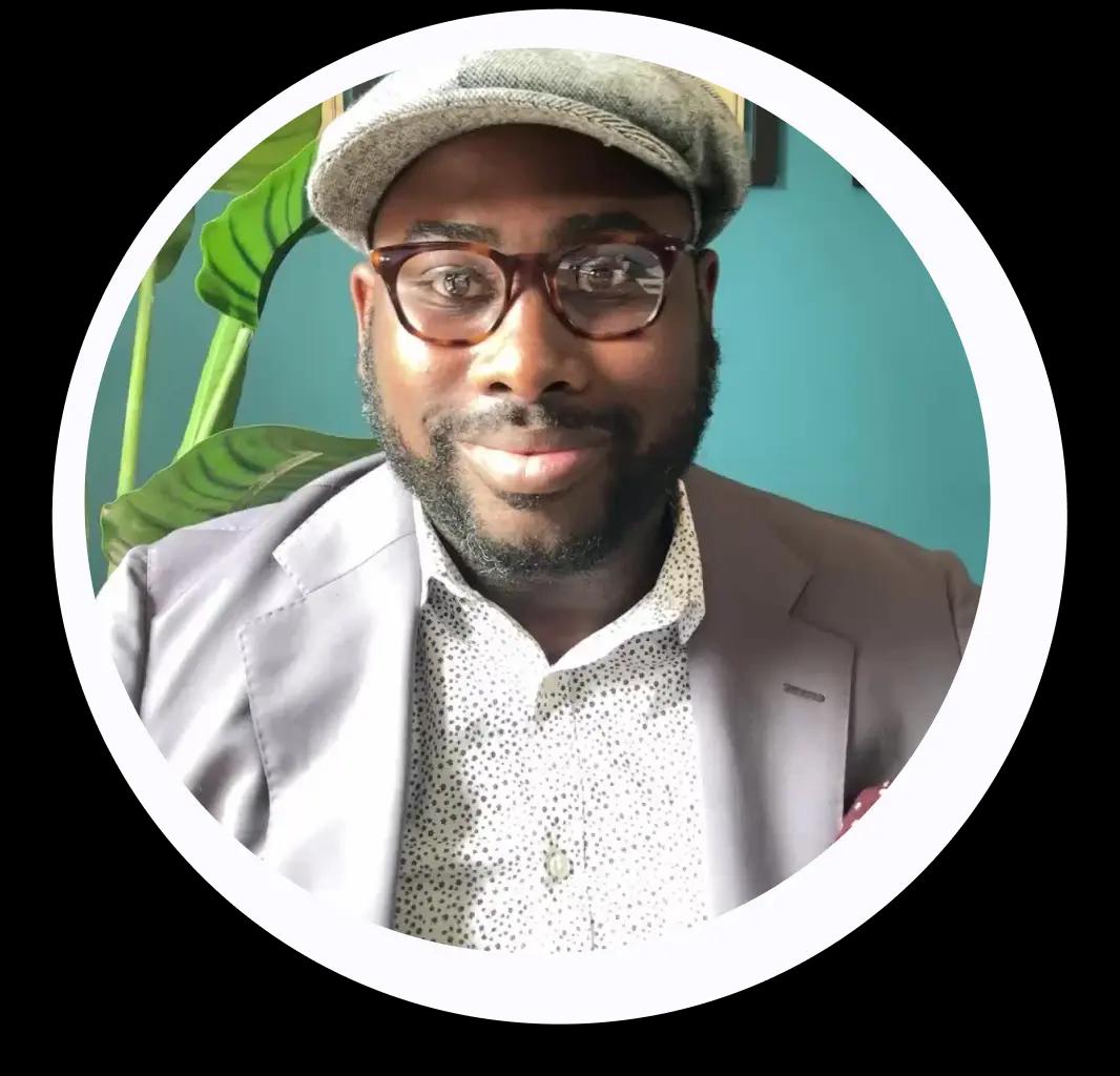 A man with glasses and a cap is smiling. He is wearing a suit and a patterned shirt, with a plant in the background.