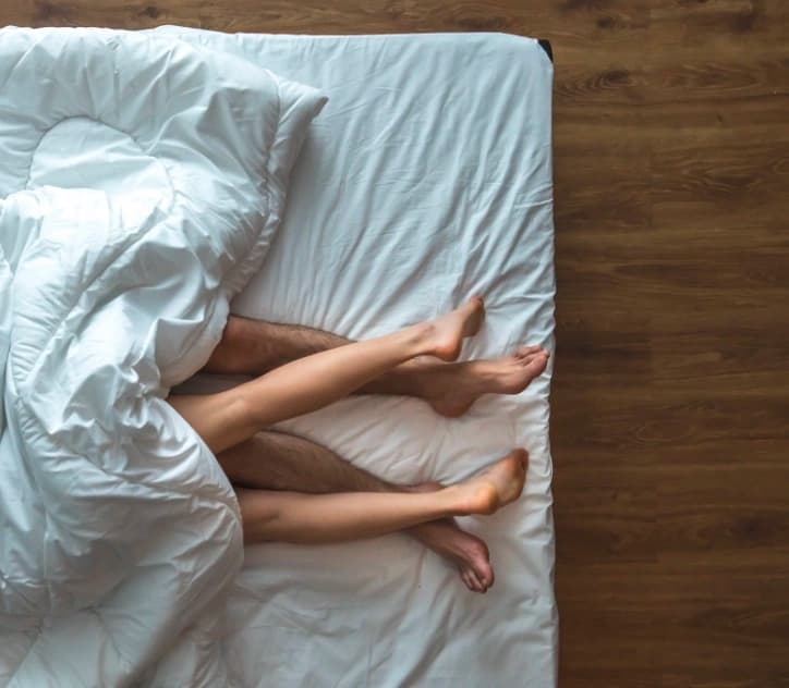 A couple is lying on a bed with white sheets, exploring questions about sex they recently googled.