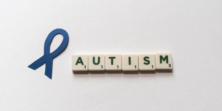 A blue awareness ribbon is placed next to Scrabble tiles spelling "AUTISM" on a white background, bringing attention to neurodevelopmental disorders.