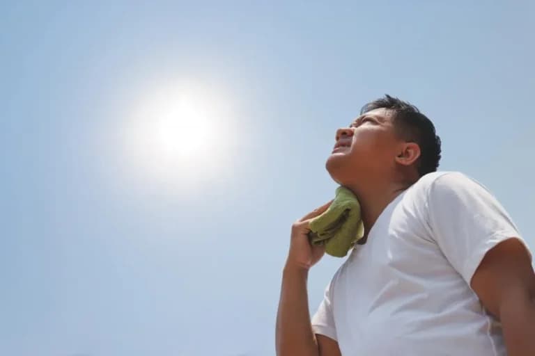 Person in a white shirt wipes their neck with a towel while standing outside under a bright sun, taking precautions against heat-related illnesses.