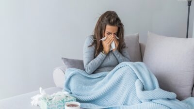 A woman is sitting on a couch, wrapped in a blanket, with a cup of coffee, battling the flu.