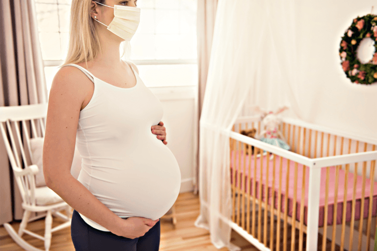 A pregnant woman wearing a surgical mask in a nursery, taking necessary precautions during the coronavirus pandemic.