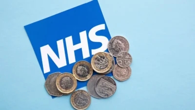 A stack of coins next to an NHS sign advertising A&E visit cost.