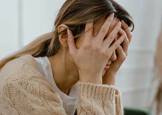 A woman with long hair and a beige sweater sits with her face in her hands, appearing distressed, possibly due to the causes of dehydration or stomach pain.