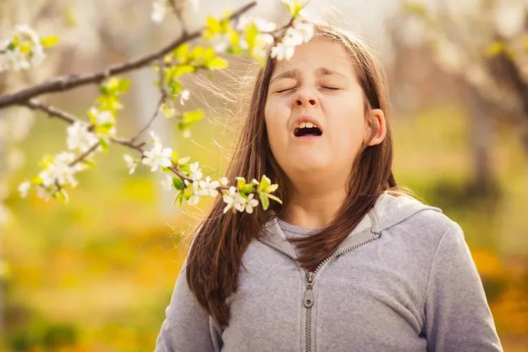 A young girl in a gray hoodie is caught mid-sneeze while standing under a blooming tree branch in a garden, clearly affected by hay fever.