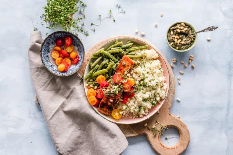 A wooden board displays a plate of quinoa, roasted vegetables, and green beans. Beside it, a bowl of cherry tomatoes and a bowl of seeds with a spoon rest on the light blue surface with a gray napkin. These foods are delicious yet considerate for those mindful of acid reflux triggers.