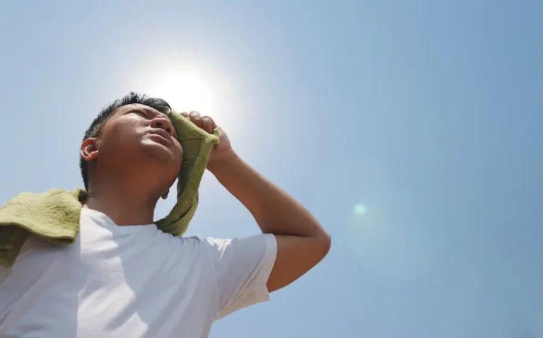A person in a white shirt wipes their forehead with a green cloth while standing under a bright sun and clear sky, possibly as a precaution against heat stroke.