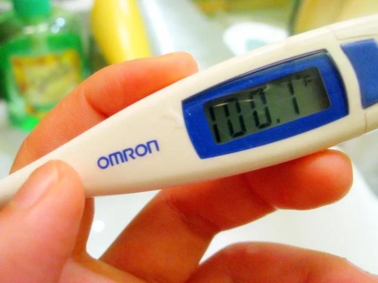 A hand holds an Omron digital thermometer displaying a fever temperature of 100.1°F.
