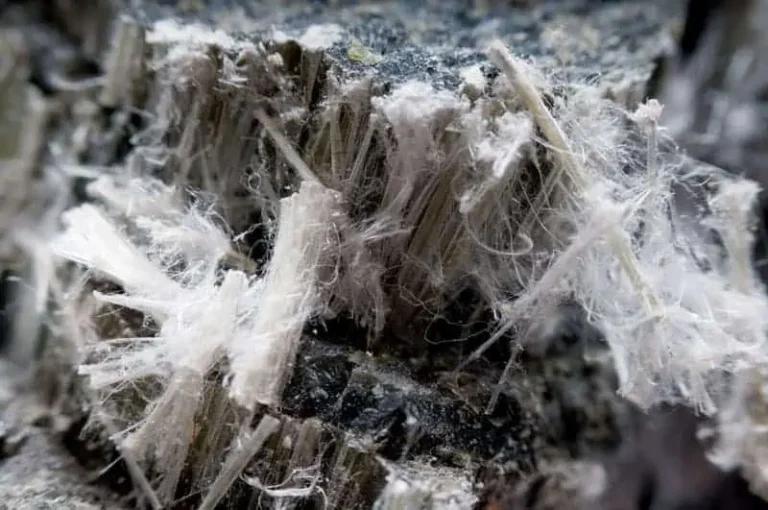 Close-up image of asbestos fibers within a rock matrix. The fibers appear in a white, fibrous, and wispy form, contrasting with the dark and rough texture of the rock. The hazardous nature of these fibers can lead to asbestosis if exposure is not managed or cured effectively.