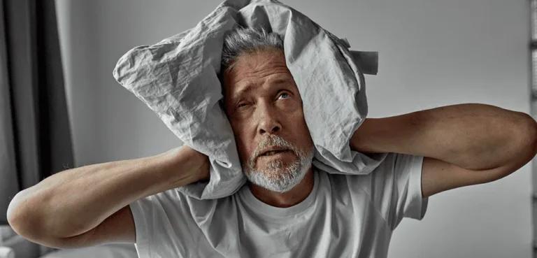 Elderly man with a distressed expression, covering his ears with a pillow, showing signs of schizophrenia