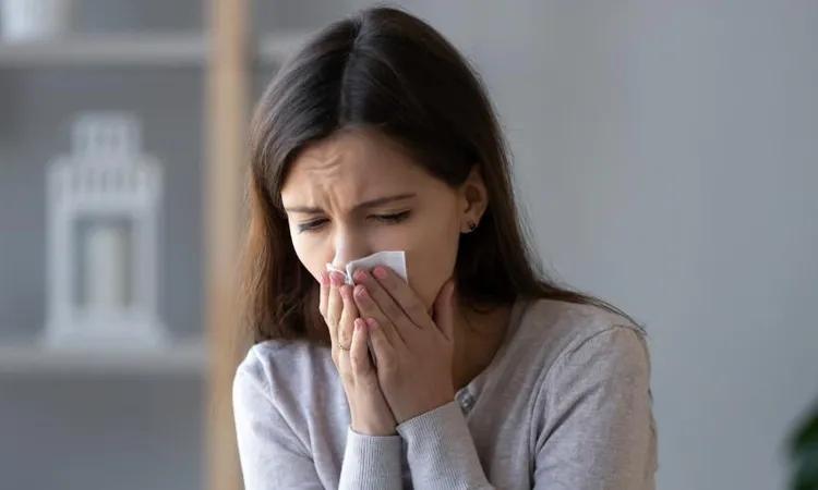 A woman with long dark hair is sneezing into a tissue, indicating a possible upper respiratory tract infection. She is wearing a light-colored sweater and is indoors.